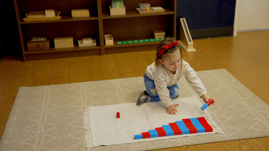 A young child in a white sweater and red headband arranges red and blue Montessori number rods on a floor mat, with shelves of Montessori materials in the background.